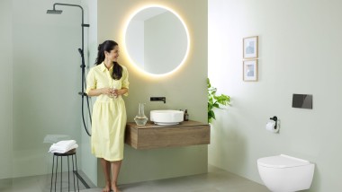 Femme en robe jaune devant une salle de bains vert menthe avec meubles et céramiques de salle de bains Geberit et robinets noirs.